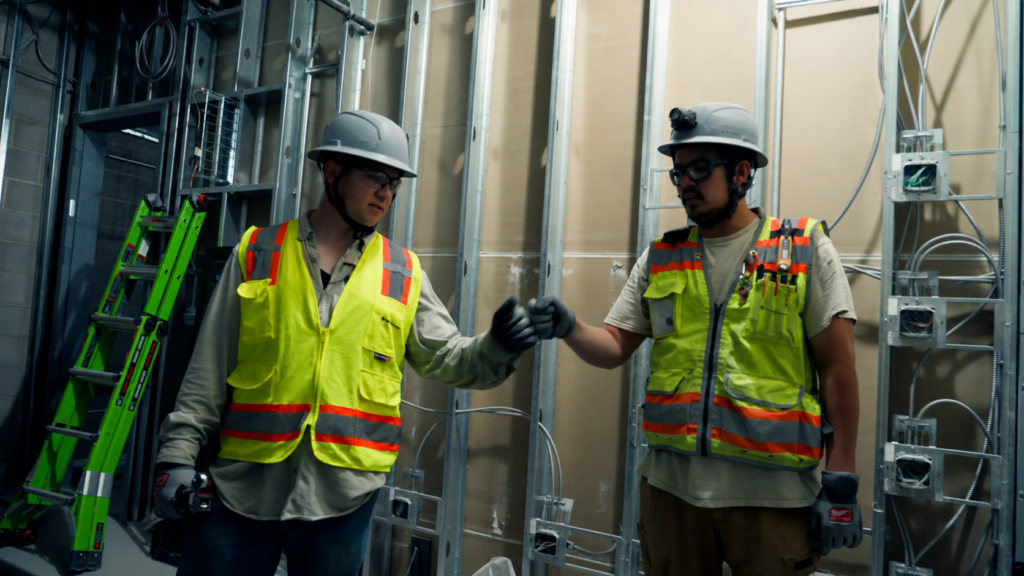 Two electricians in hard hats and safety vests fist-bump beside a framed wall of boxes and conduit